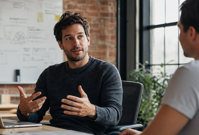 Two coworkers discuss ideas at a wooden desk in a bright open-office, with one man gesturing as he speaks.